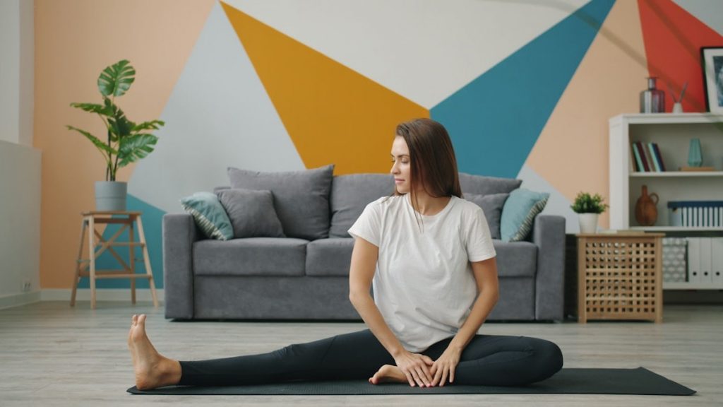 Attractive girl in leggings and T-shirt is doing stretching exercises on yoga mat relaxing on floor at home busy with practise. Youth, lifestyle and health concept.